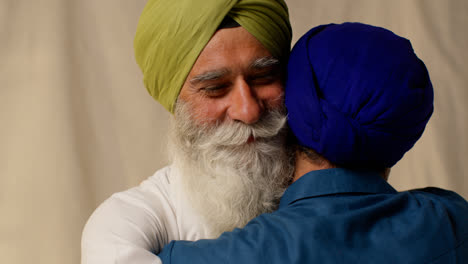 Close-Up-Studio-Shot-Of-Senior-Sikh-Father-Embracing-Adult-Son-Both-Wearing-Turbans-Against-Plain-Background